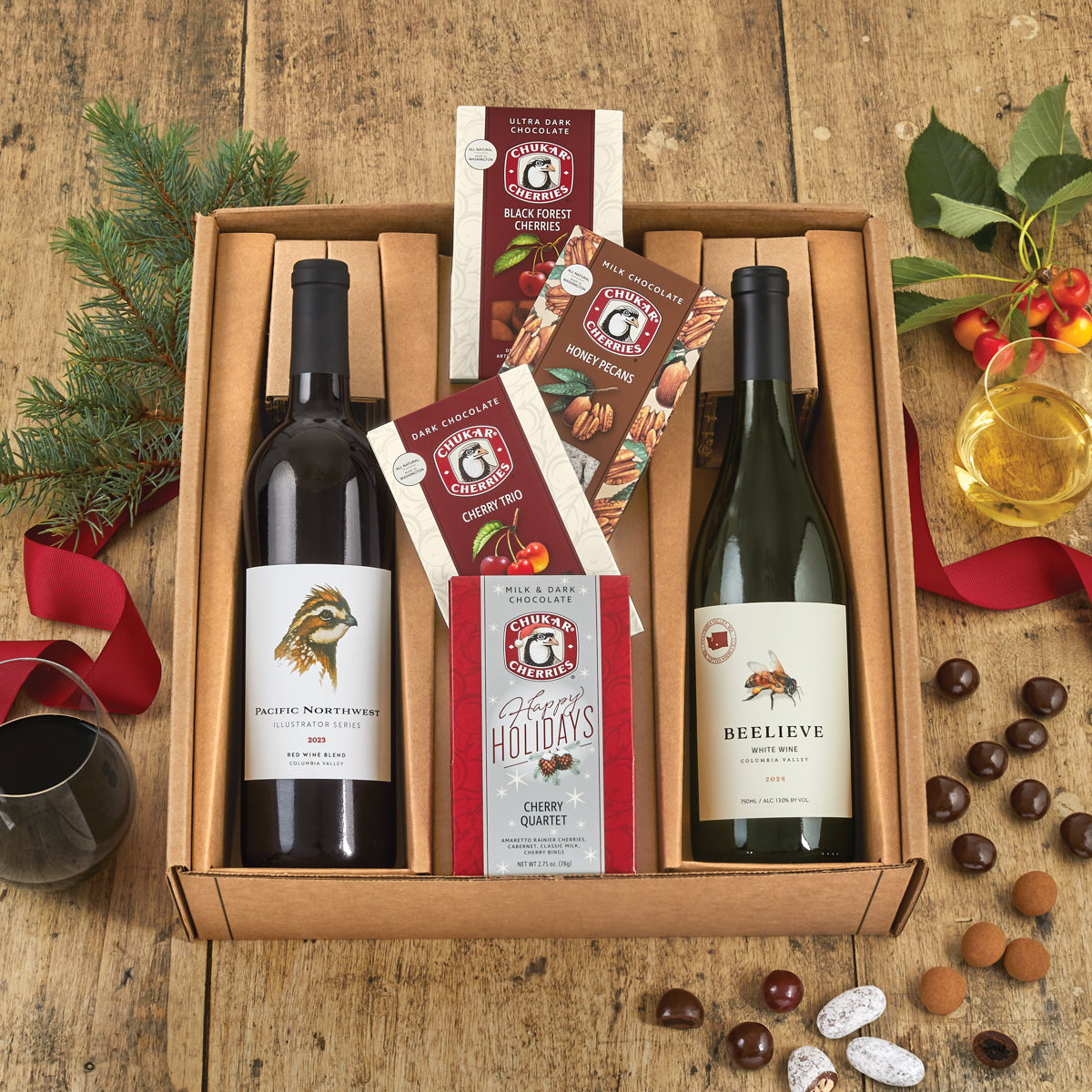 Open gift box of chukar cherries wines and chocolates, a glass of each wine, and spills of cut and whole chocolates