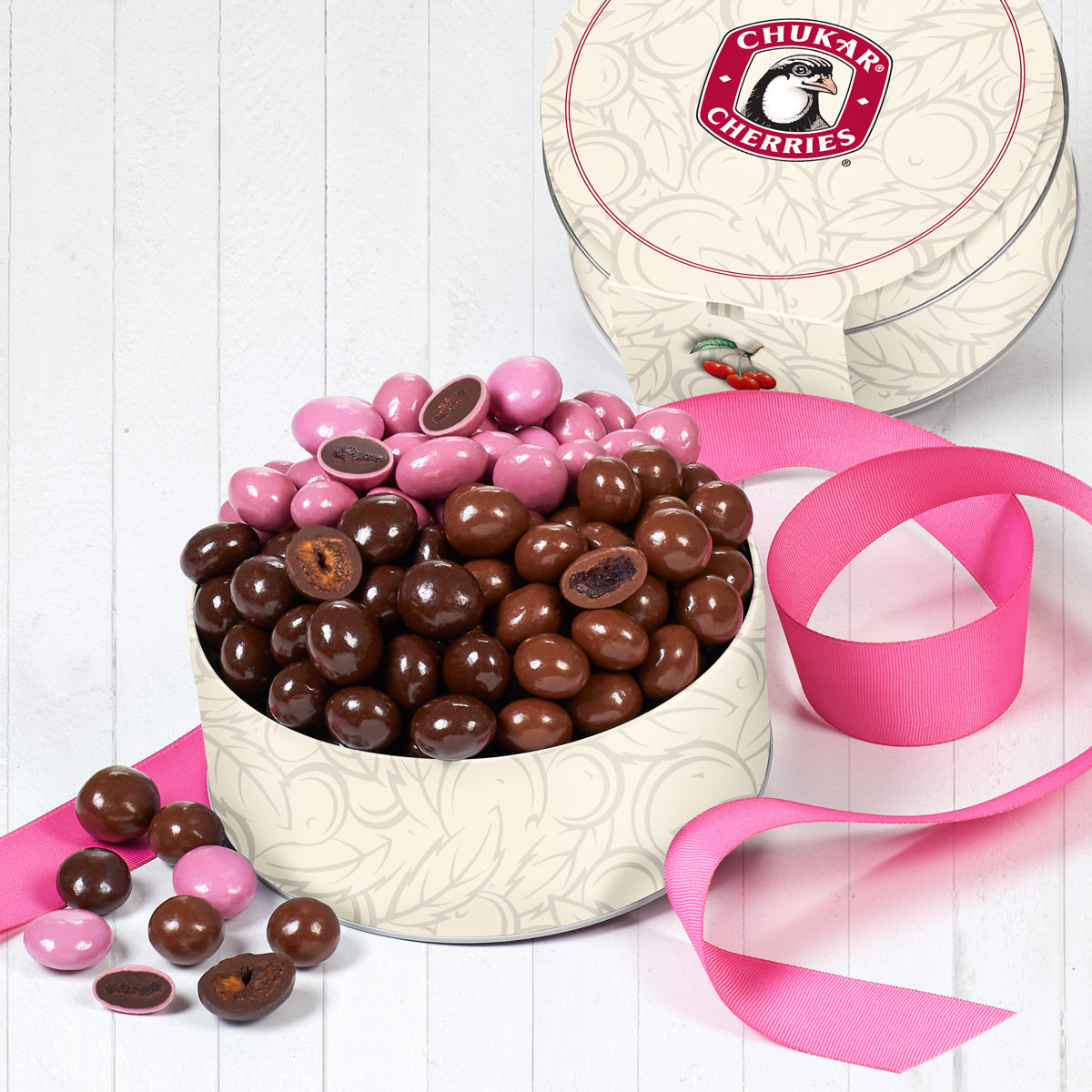 Chocolate covered cherries in a cream tin encircled by a pink ribbon on a white background.