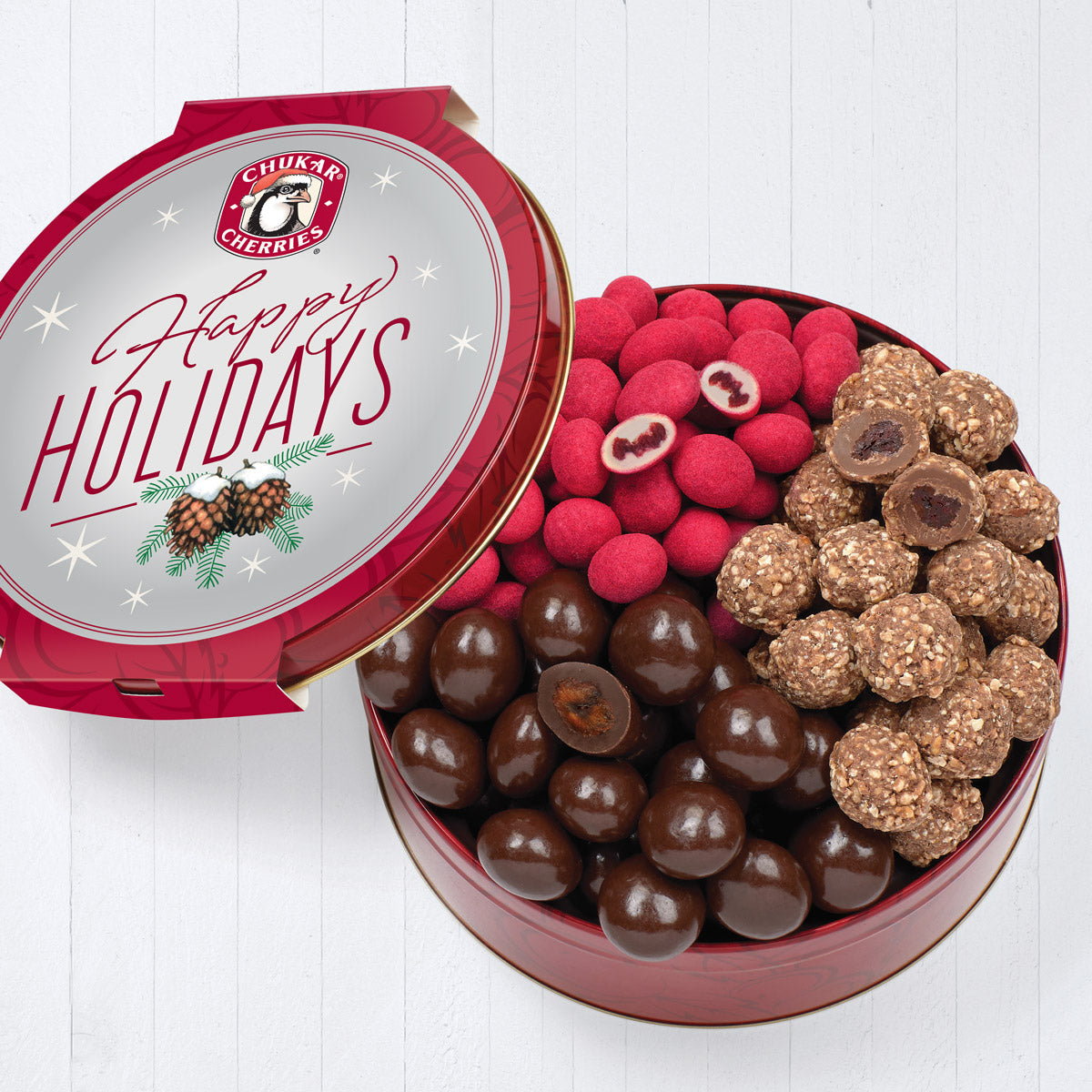 Happy holidays trio tin shown with lid off with cut and whole white chocolate raspberry truffles, milk chocolate hazelnut cherries, and classic dark chocolate cherries 