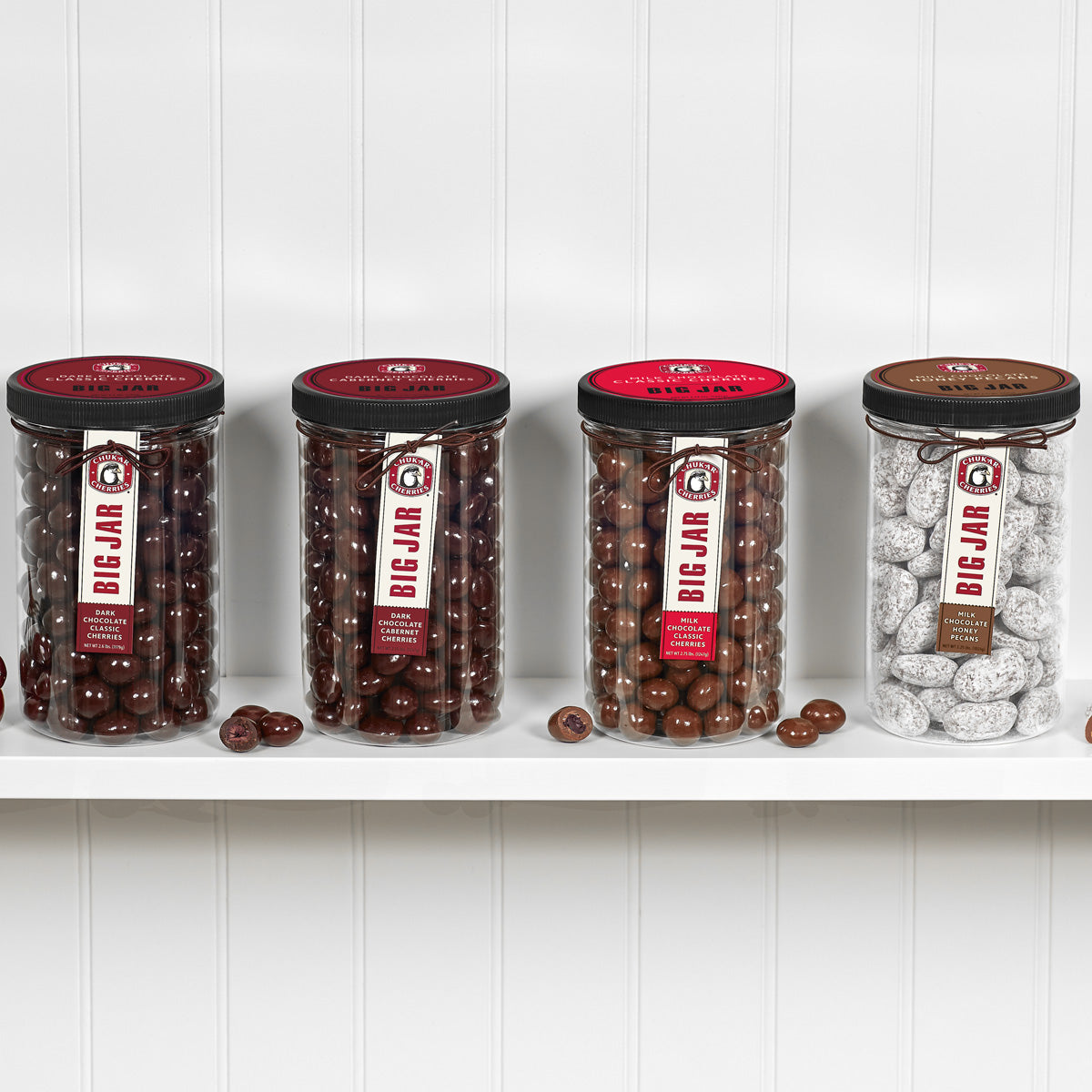 Four closed chocolate cherry and nut big jars and cut chocolates on a shelf. In order from left, big jars are dark chocolate classic cherries, dark chocolate cabernet cherries, milk chocolate classic cherries, and milk chocolate honey pecans.  