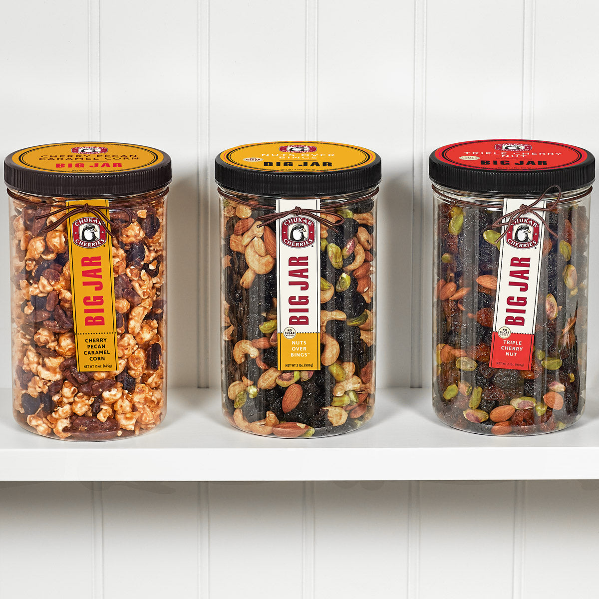Three closed cherry and nut big jars on a shelf. In order from left, big jars are cherry pecan caramel corn, no sugar added nuts over bings, and no sugar added triple cherry nut. 