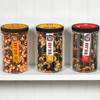 Three closed cherry and nut big jars on a shelf. In order from left, big jars are cherry pecan caramel corn, no sugar added nuts over bings, and no sugar added triple cherry nut. 