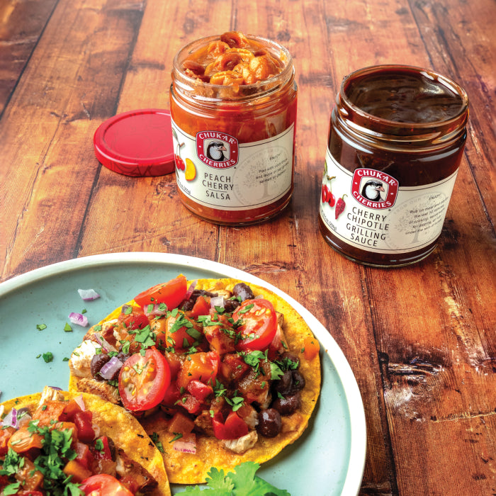 Jars of peach cherry salsa and cherry chipotle grilling sauce on a wood table with a plate of tostadas.