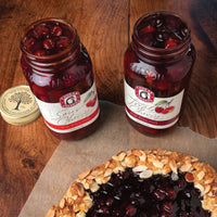 Open jars of Sour Cherry Pie Filling and Triple Cherry Pie Filling, adjacent to a rustic tart on a wooden kitchen table